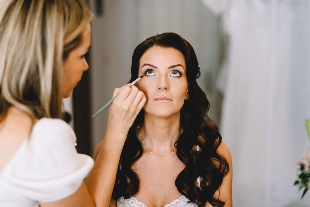 Makeup artist applying eye makeup on a bride with dark wavy hair wearing a strapless wedding dress.