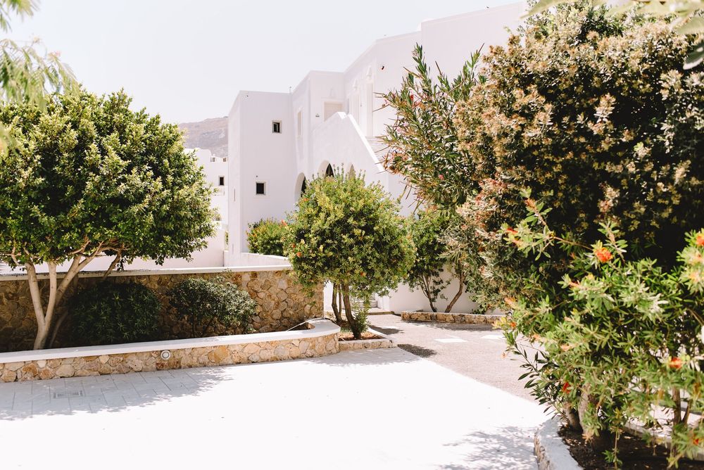 White Mediterranean building surrounded by lush green trees and stone wall courtyard in bright sunlight.