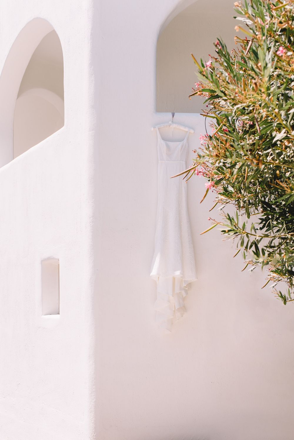 White wedding dress hanging on a hanger against a whitewashed wall with arches and green flowering tree in Santorini.