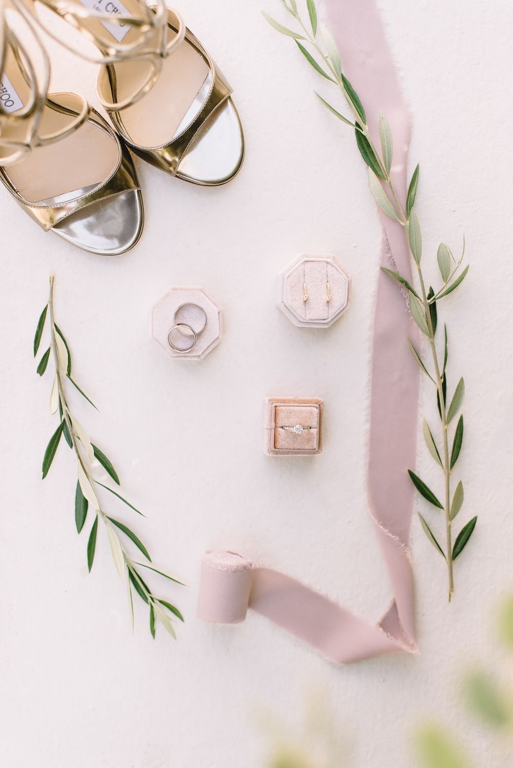 Gold bridal heels, pink velvet ring boxes with diamond rings, drop earrings, and olive branch greenery with blush ribbon flatlay.