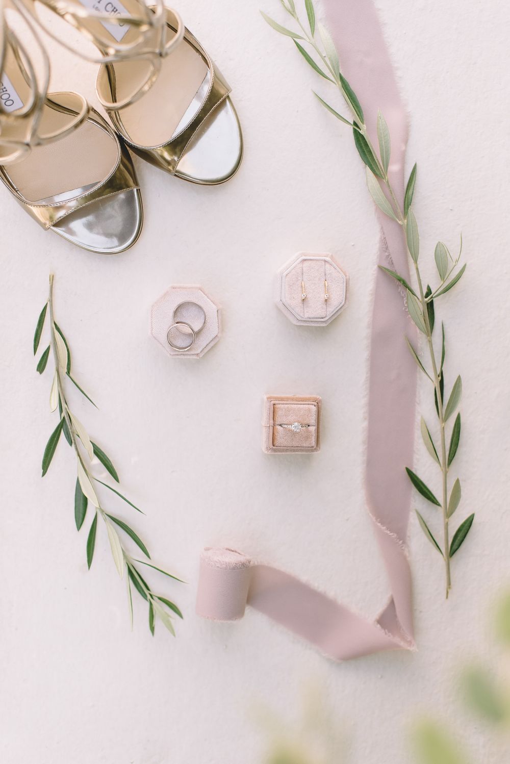 Elegant wedding accessories including gold shoes, ring boxes and delicate olive branches arranged on light surface.
