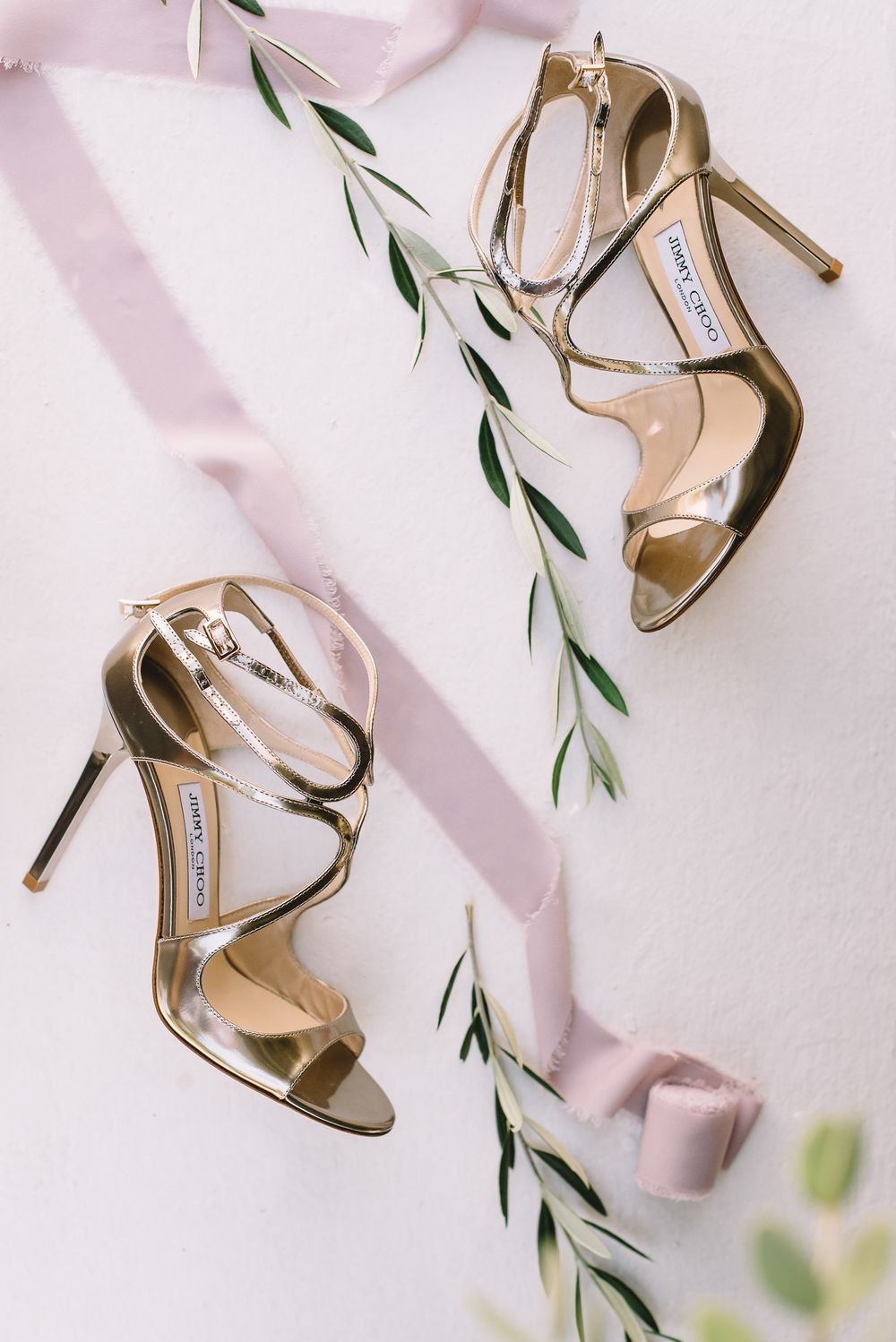 Gold Jimmy Choo strappy heeled sandals displayed with pink ribbon and olive branches on white background.