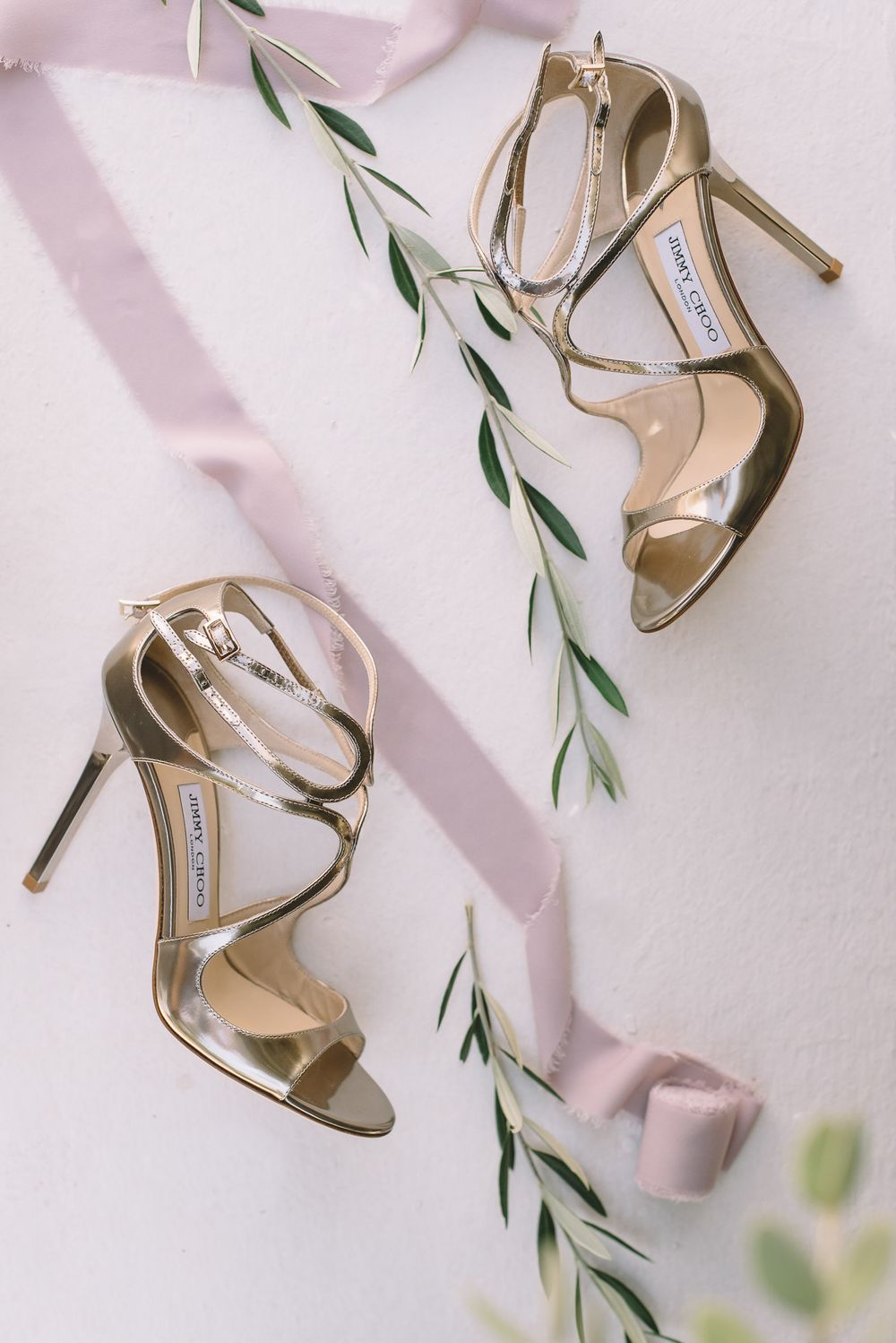 Elegant gold Jimmy Choo bridal heels with delicate straps styled with pink ribbon and eucalyptus leaves.