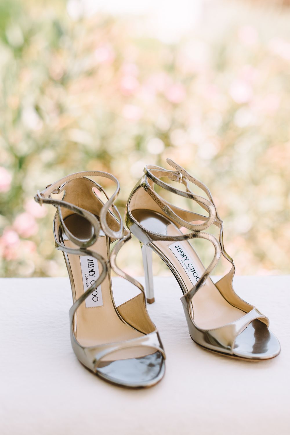 Silver Jimmy Choo strappy high heel sandals displayed on a white surface with soft floral bokeh background.