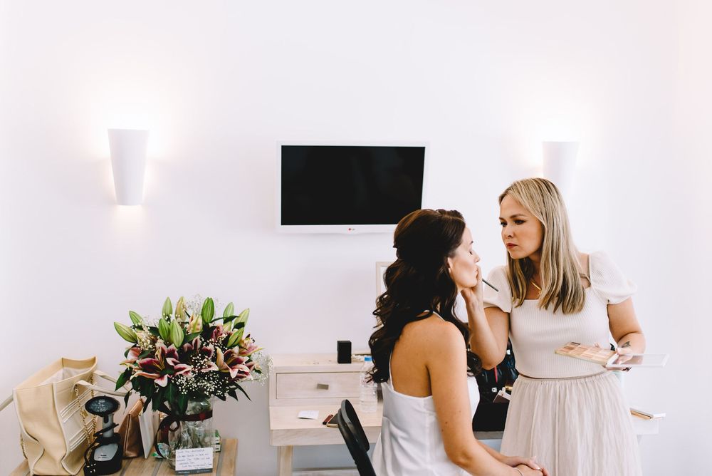 Makeup artist applying makeup to a bride in a bright white room with floral arrangements nearby.