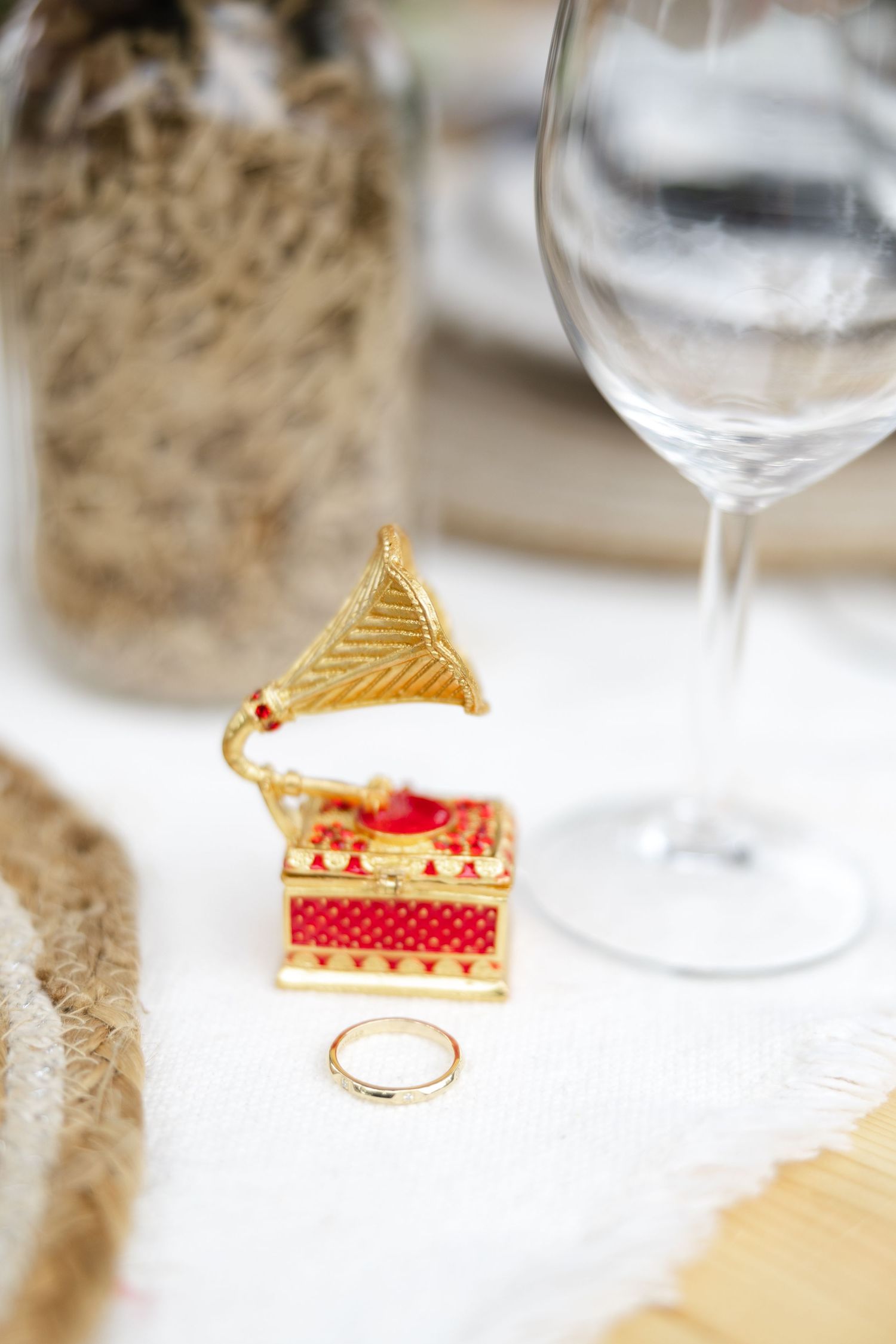 Gold decorative gramophone ring box and gold engagement ring on white table beside wine glass with textured cloth decor.