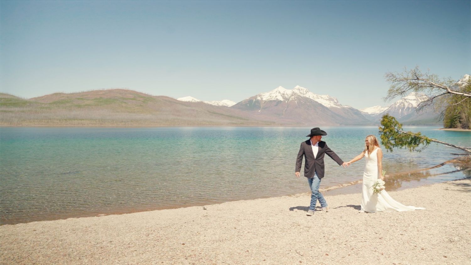 A couple walks hand in hand along a sandy lakeside beach with snow-capped mountains and turquoise waters in the background.
