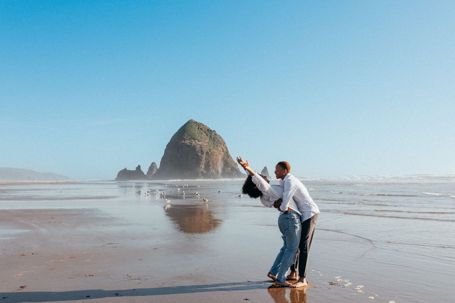 A couple embrace on the beach at Cannon Beach, Oregon with Haystack Rock rising prominently in the background.