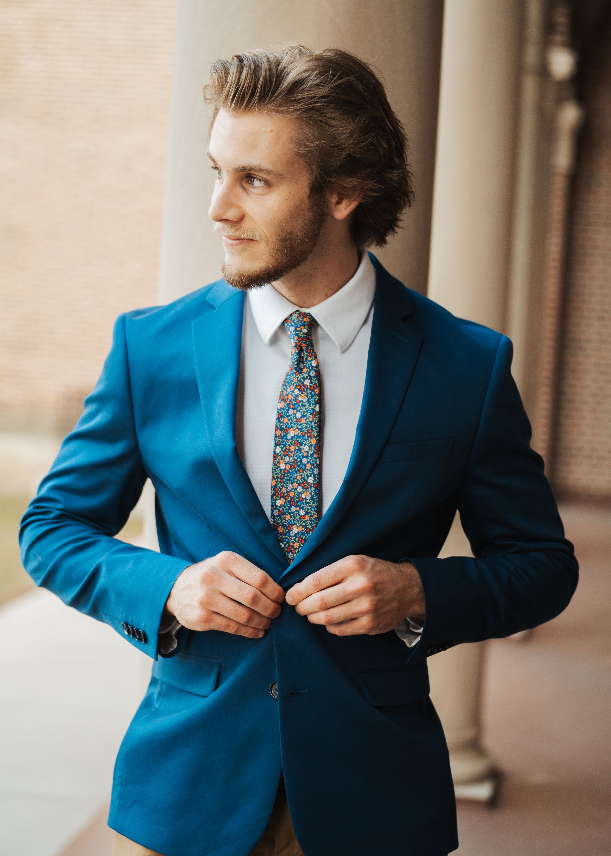 Ethan adjusting his navy suit and floral tie during a headshot session, standing in front of marble pillars.