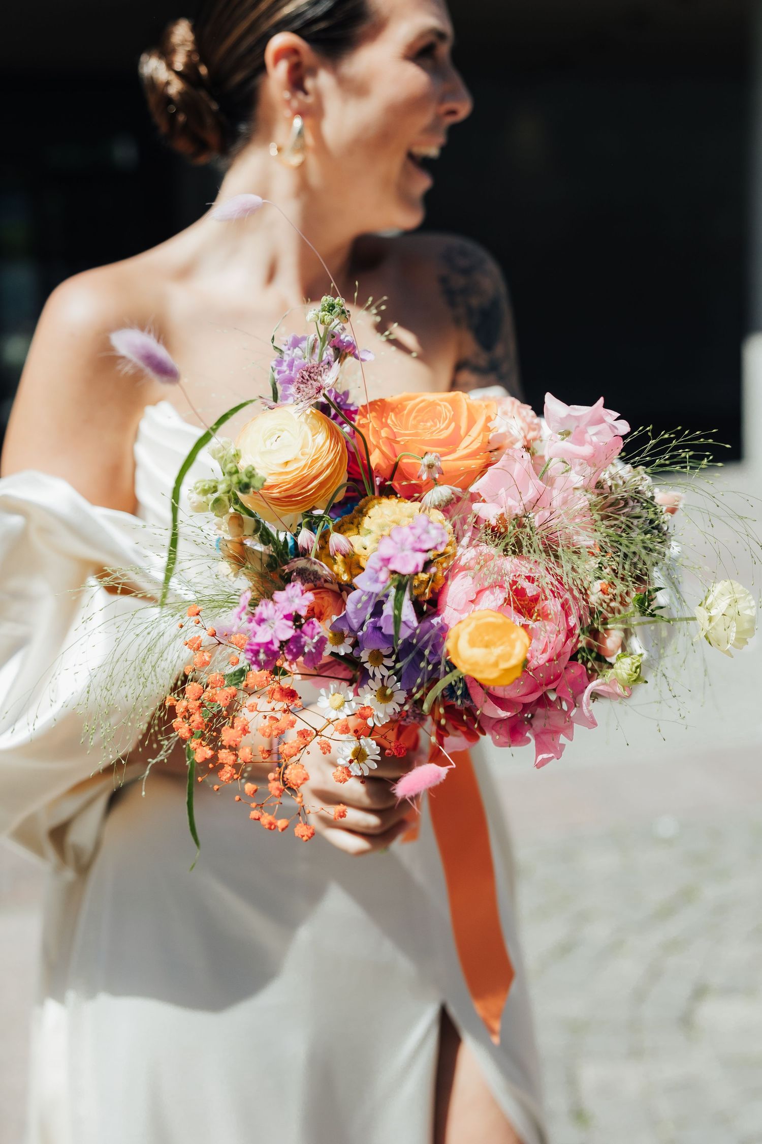 A bright and colorful bridal bouquet with orange ranunculus, pink and purple flowers, held against a white wedding dress.