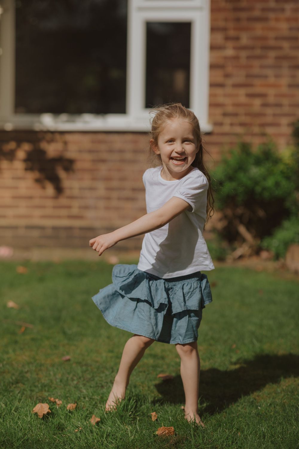 A child spins and plays joyfully on a sunny lawn with autumn leaves near a brick house.