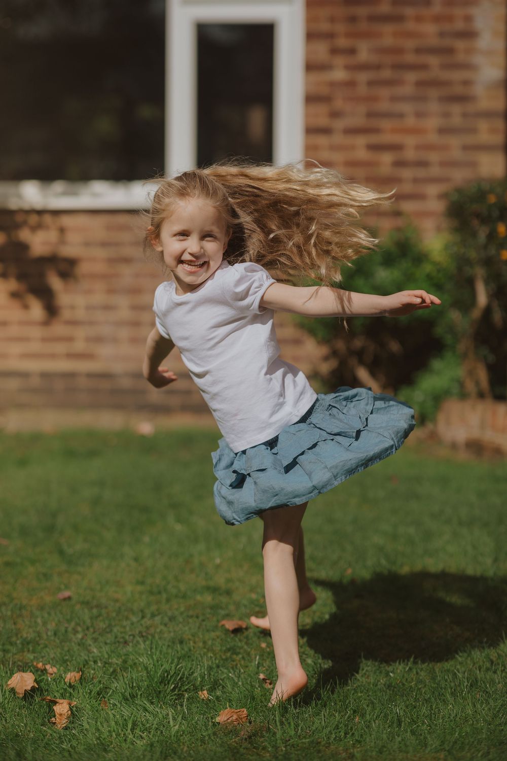 Energetic child leaps and twirls on a grassy lawn in front of brick house in white shirt and denim.