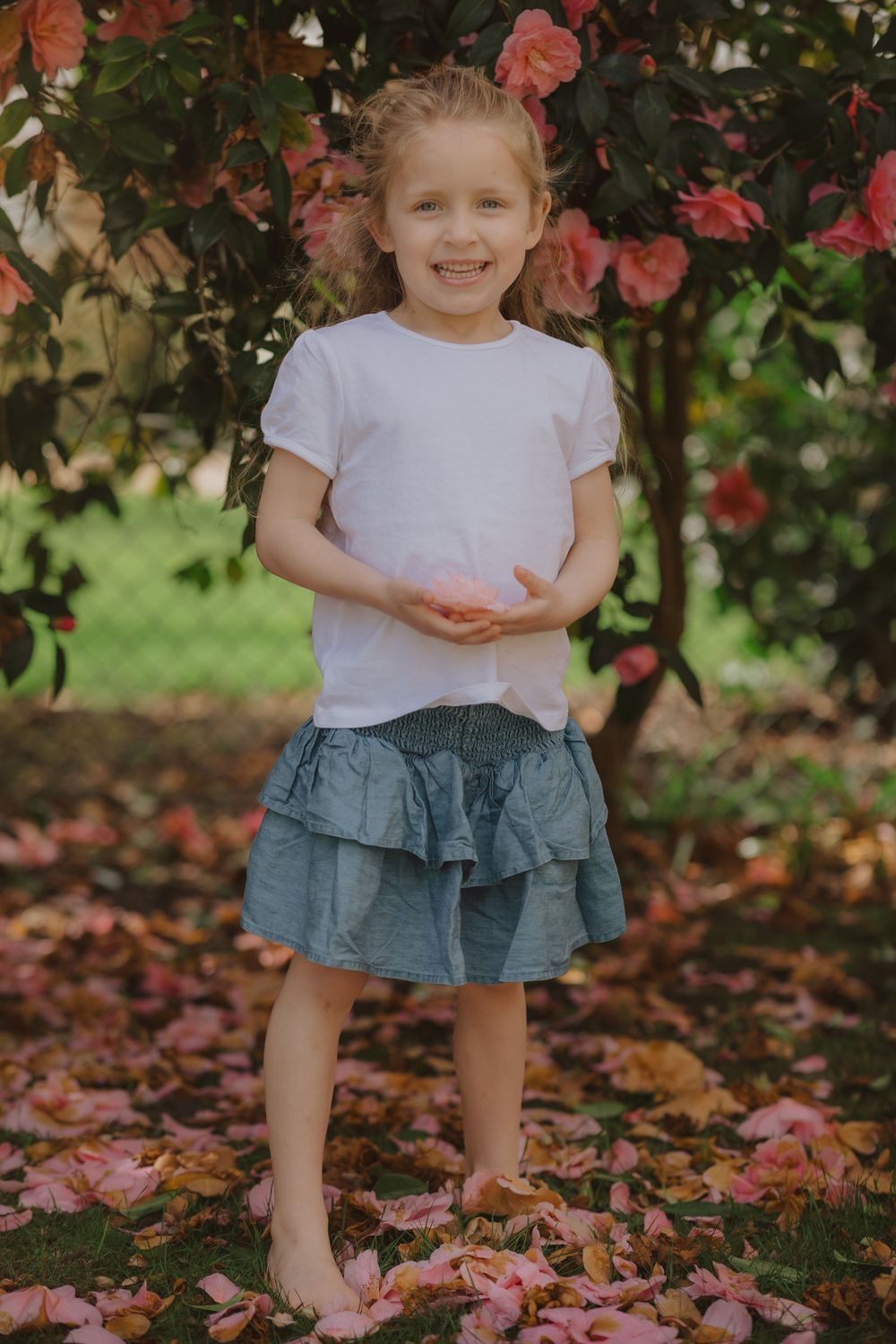Child standing in a white shirt and blue ruffle skirt beneath pink flowering branches with fallen petals.