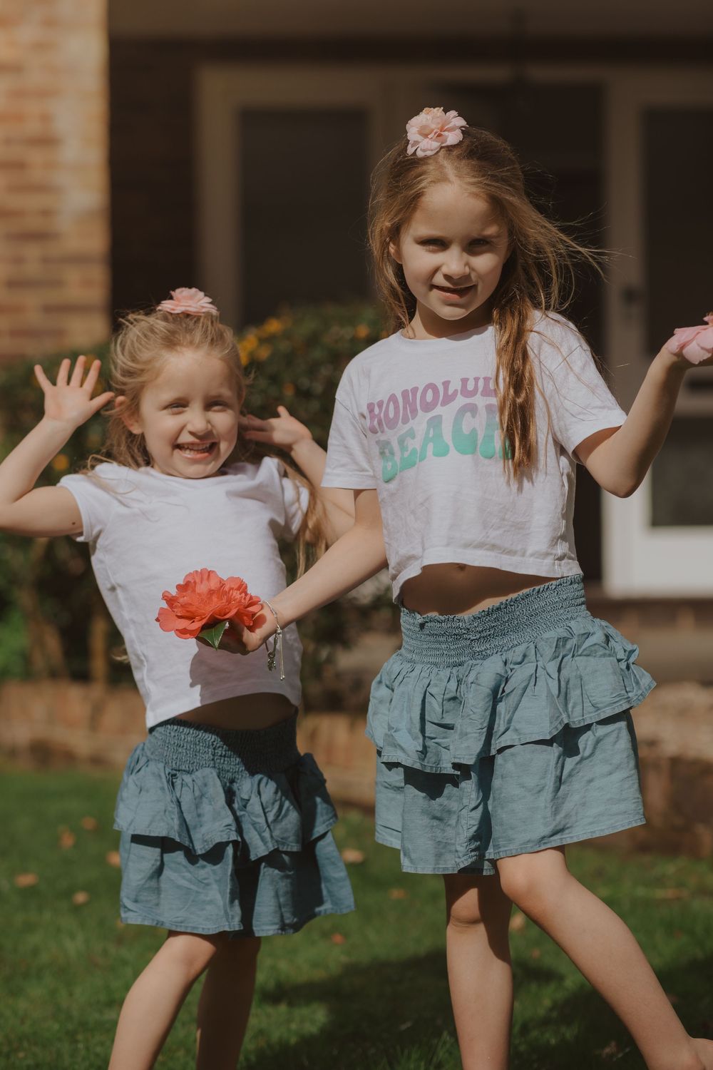 Two children in matching denim ruffle skirts dancing outdoors with pink flowers.