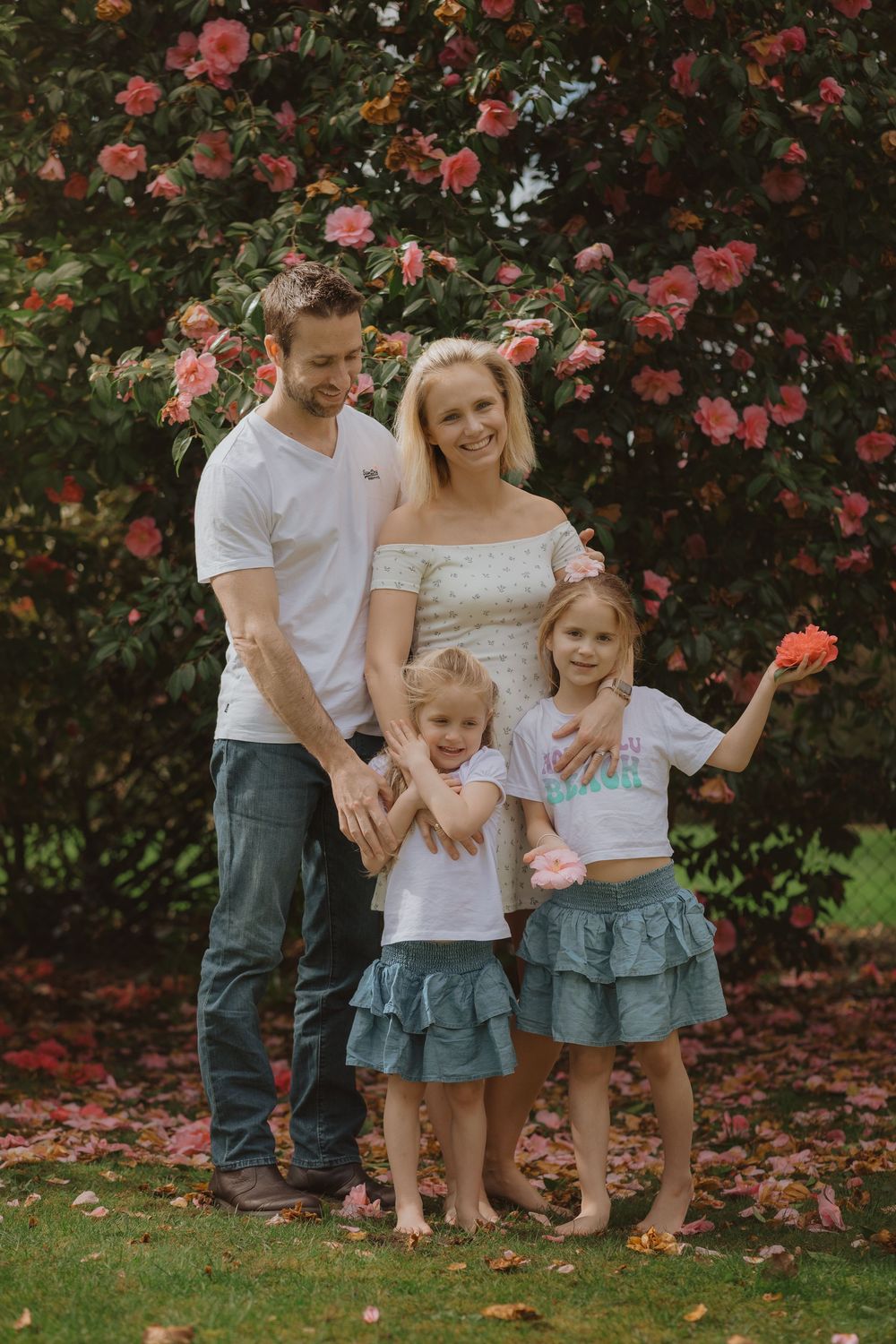 Family portrait under blooming coral camellia tree with fallen petals on grass.