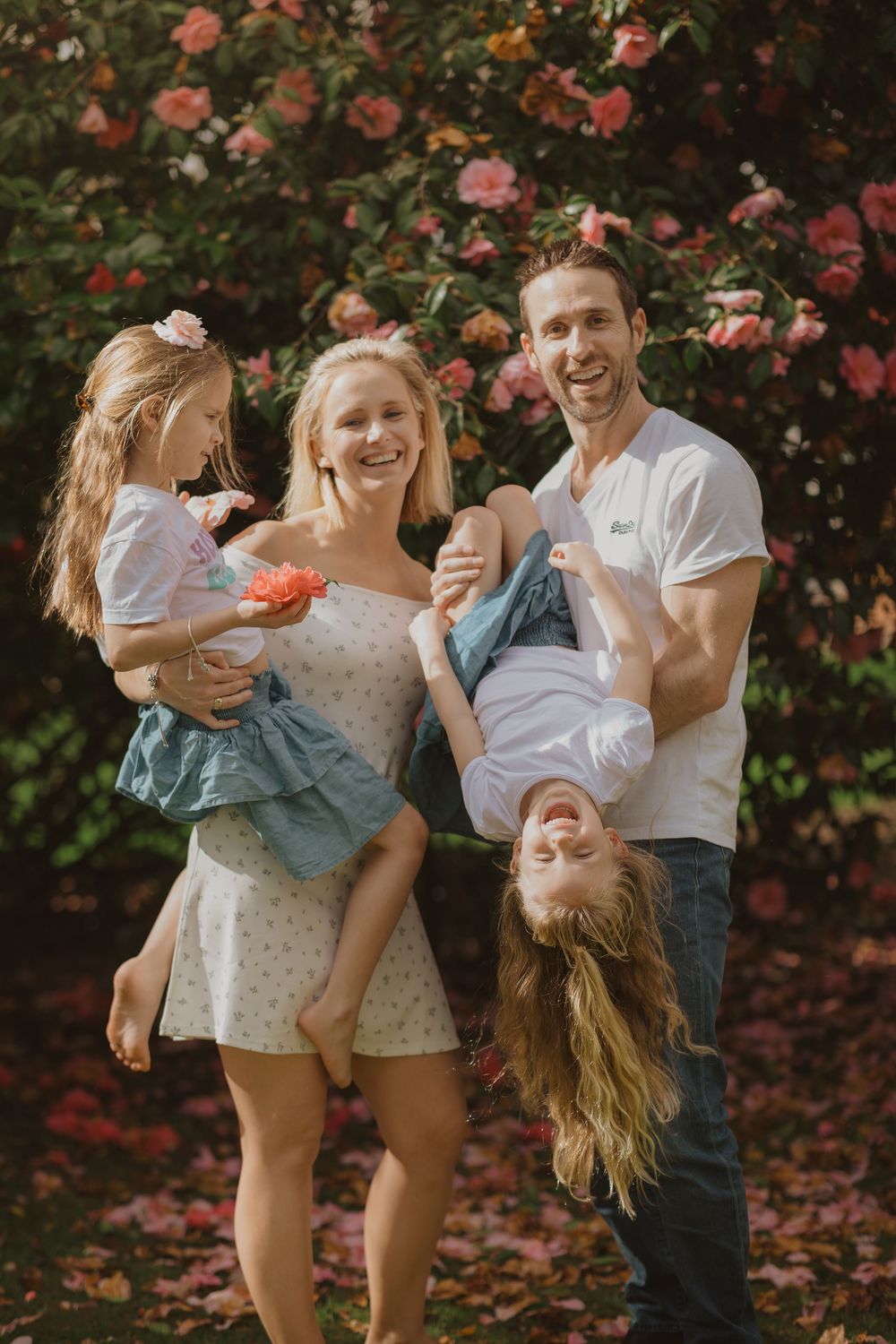 Family plays and laughs together in front of blooming pink rose bushes on a sunny day.