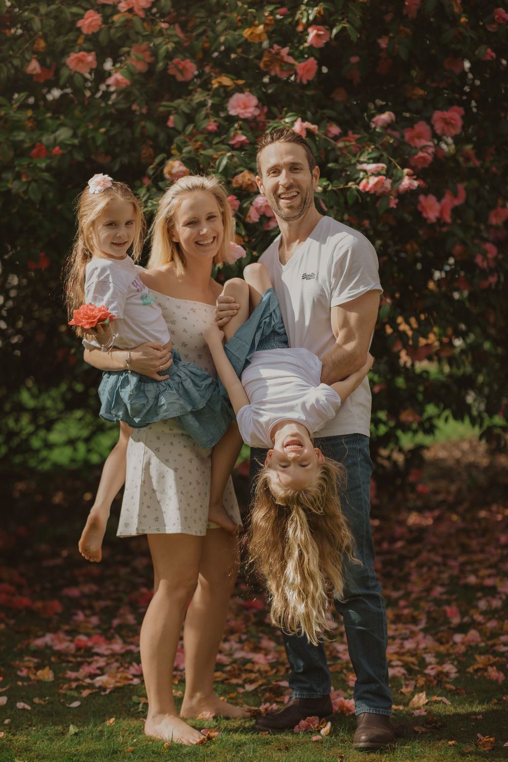 A playful family shares laughter and embraces while standing together against blooming pink flowers in a garden.