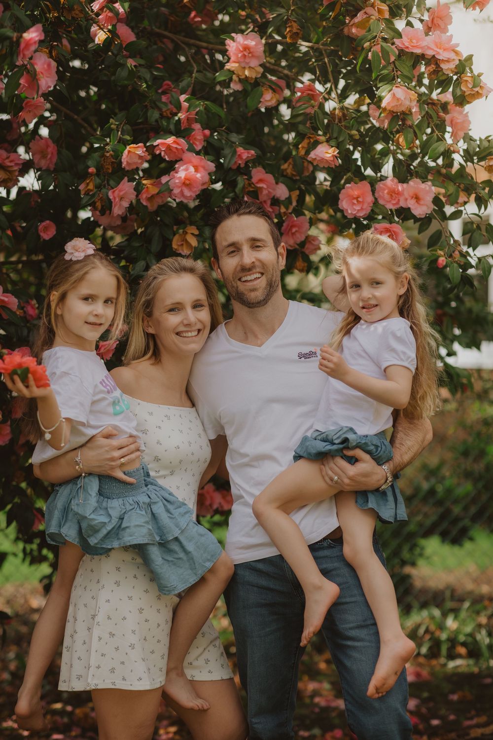 Family poses for portrait in front of pink flowering bushes, all wearing casual white and blue outfits.