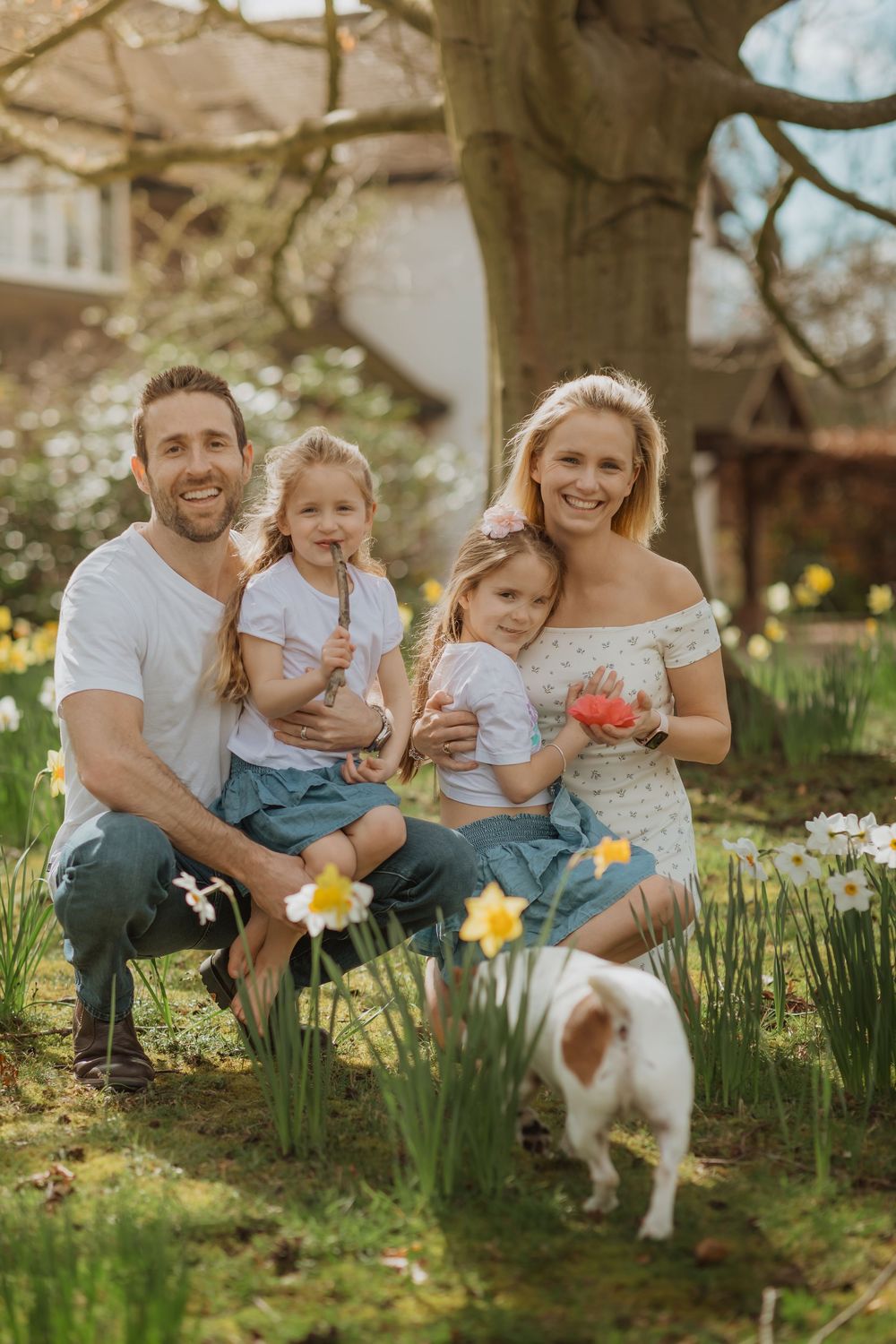Family poses together in spring garden with blooming daffodils and small dog in natural outdoor setting.