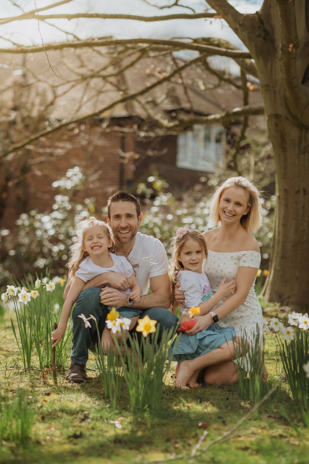 Family outdoor photoshoot in garden with blooming spring flowers and brick house background.