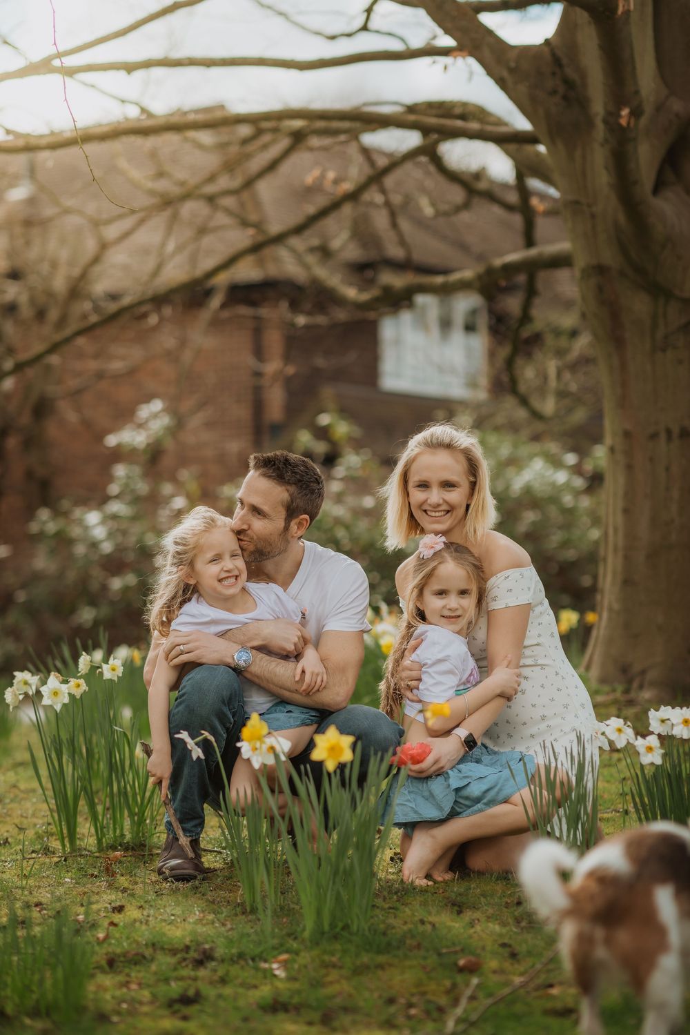 Candid family moment in garden with daffodils and small pet dog visible in corner.