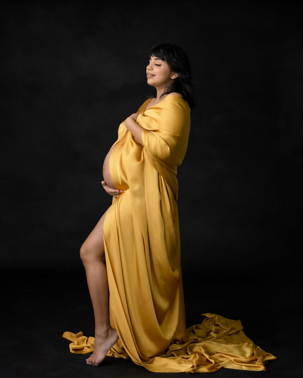 Dramatic maternity pose in flowing mustard yellow gown against black background.
