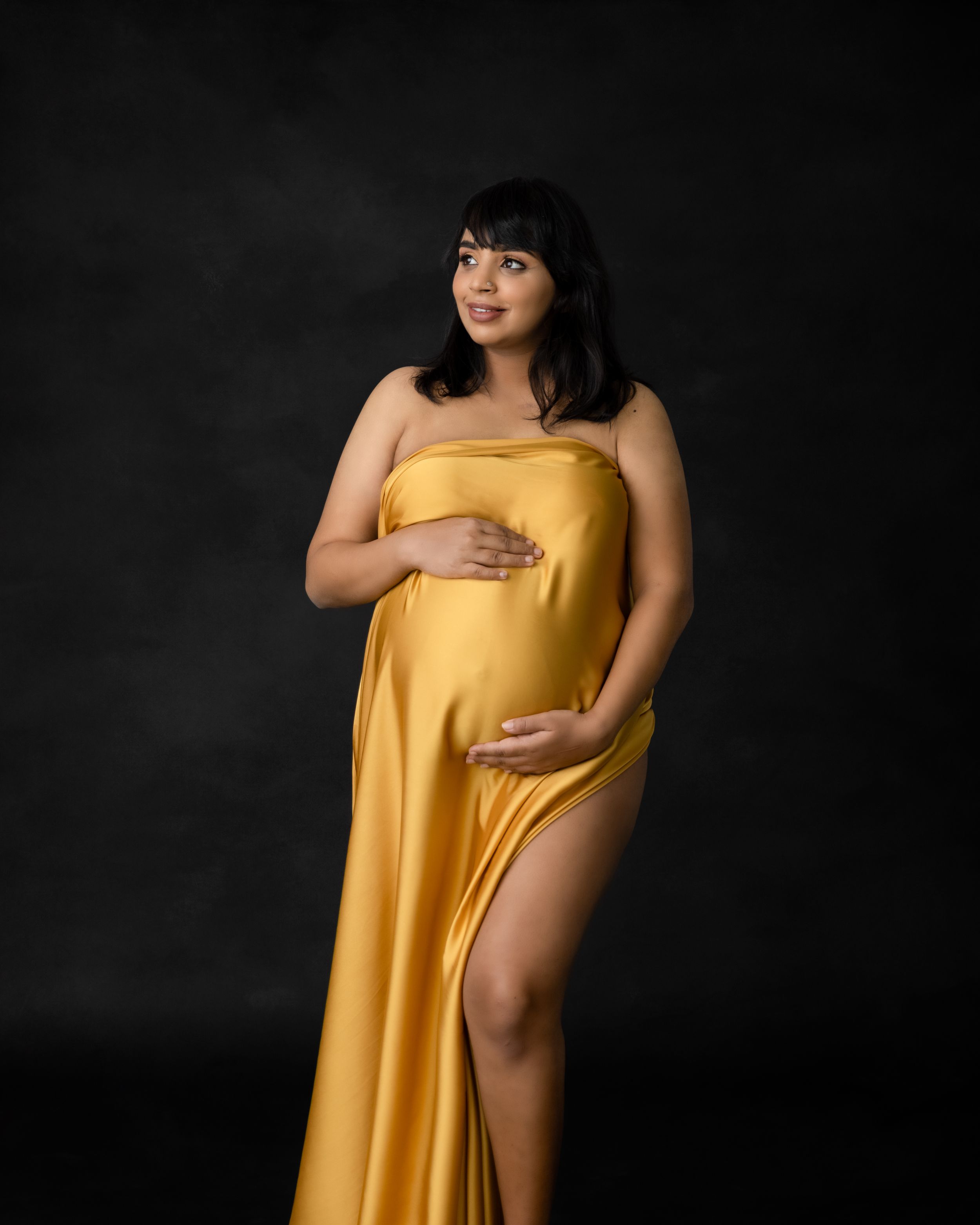 Elegant maternity pose in form-fitting golden dress with thigh slit against dark backdrop.