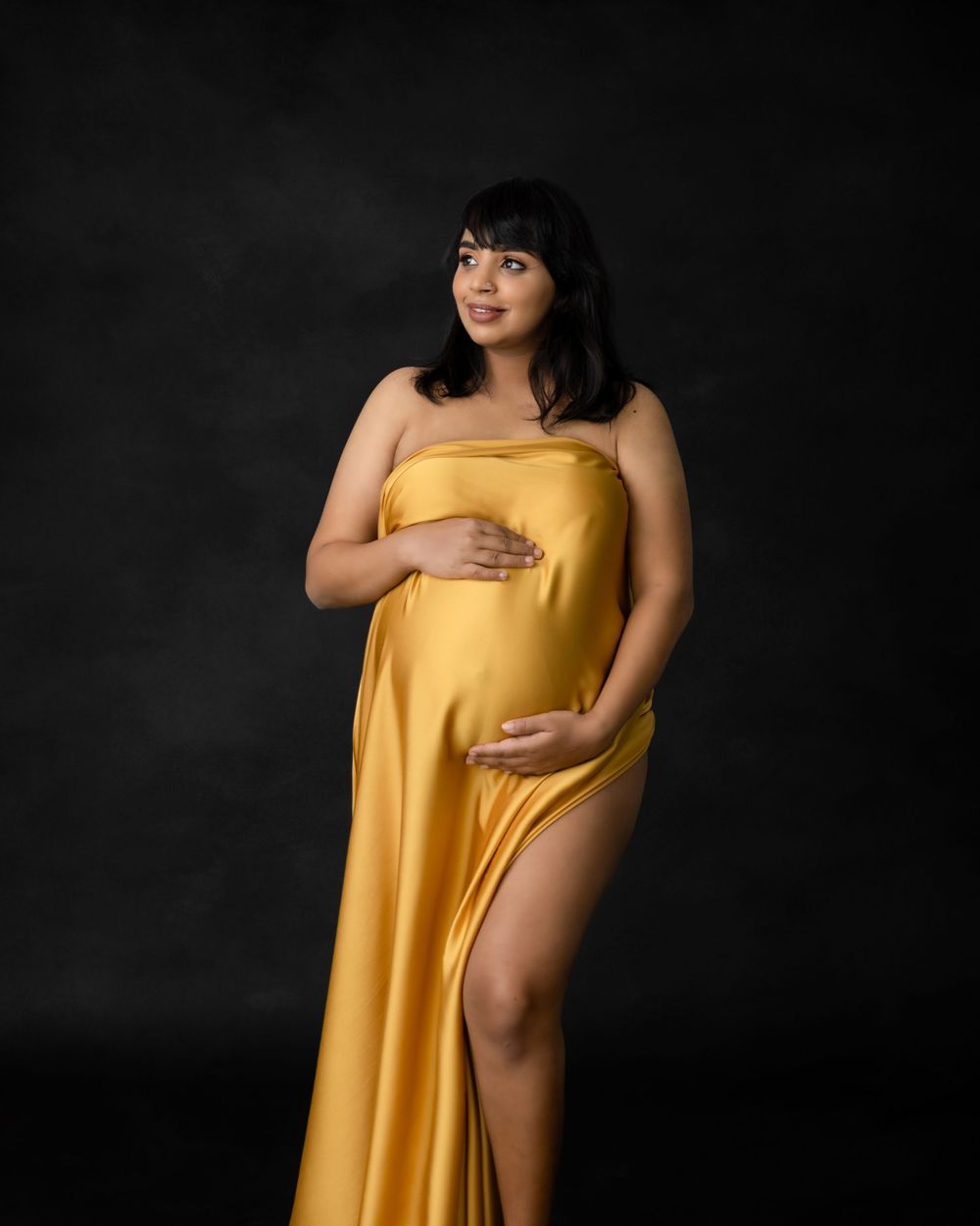 Elegant maternity pose in form-fitting golden dress with thigh slit against dark backdrop.
