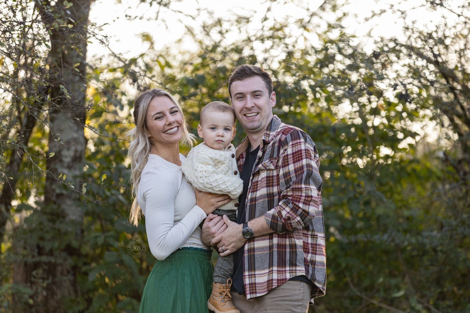 A family stands together outdoors during golden hour in a natural setting with trees and greenery in the background.