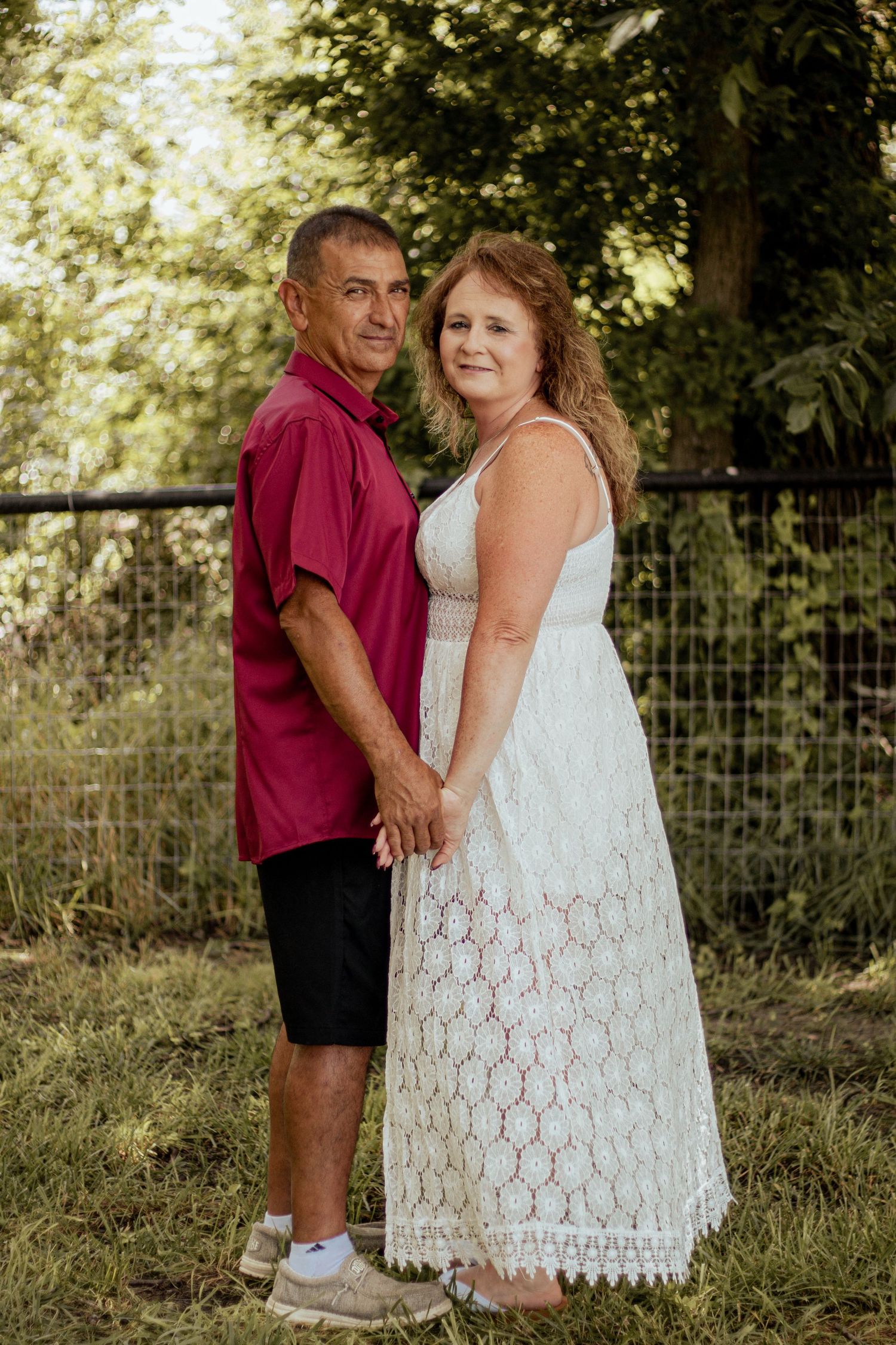 A couple poses outdoors in summer attire, with one in a red shirt and shorts and the other in a white lace dress.