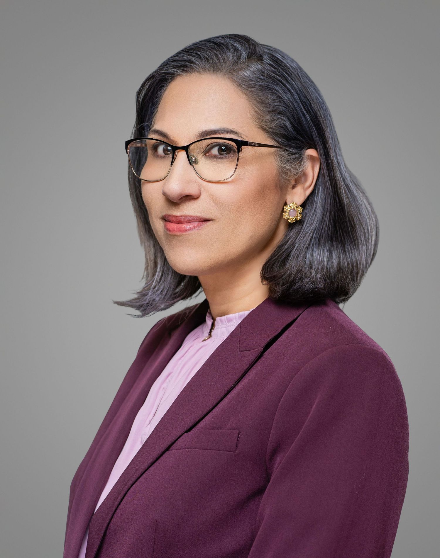 Professional portrait of a businesswoman wearing glasses and burgundy blazer with a friendly expression.