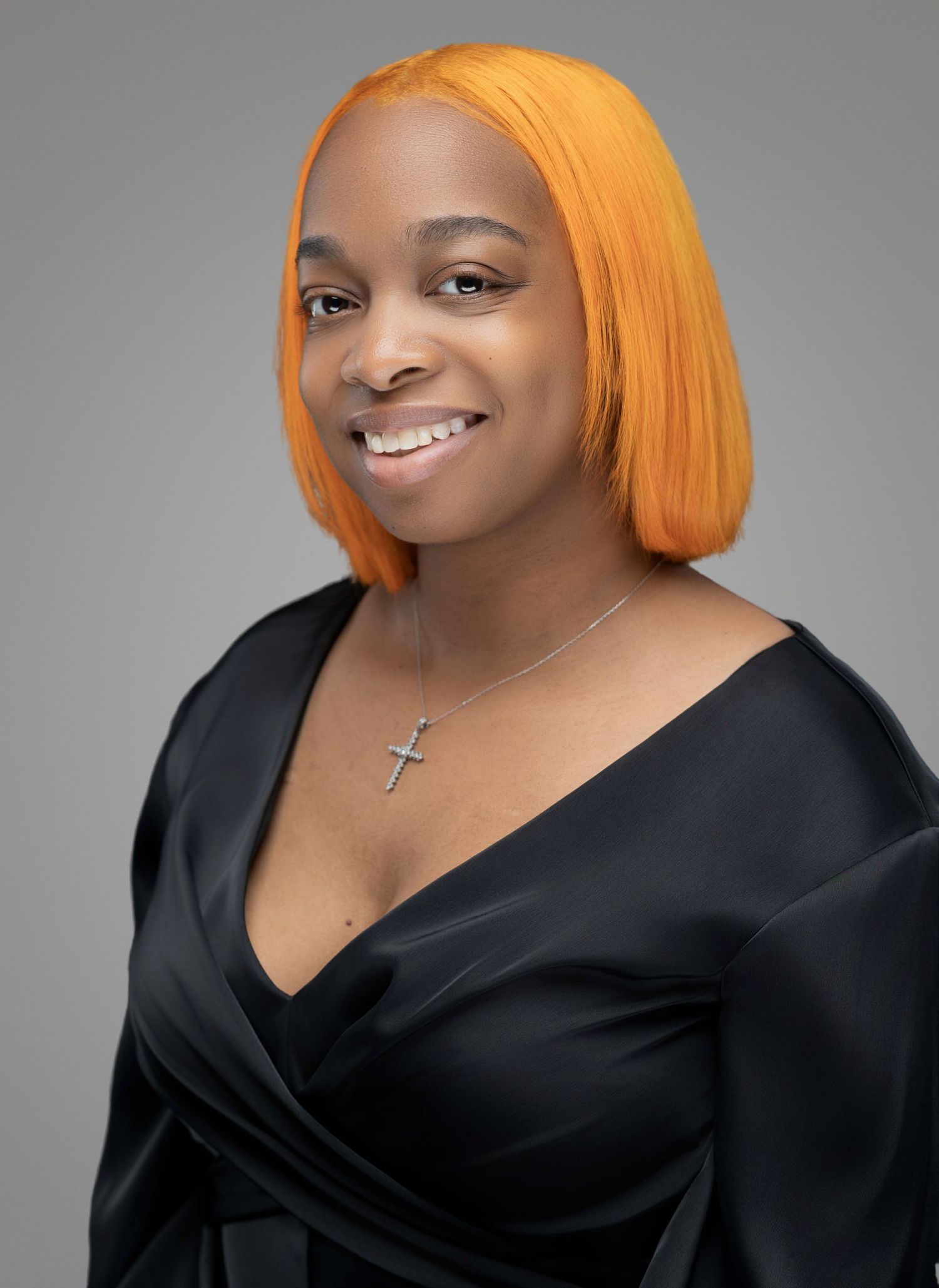 Professional studio portrait of someone with vibrant orange hair wearing a black wrap dress and cross necklace against gray backdrop.
