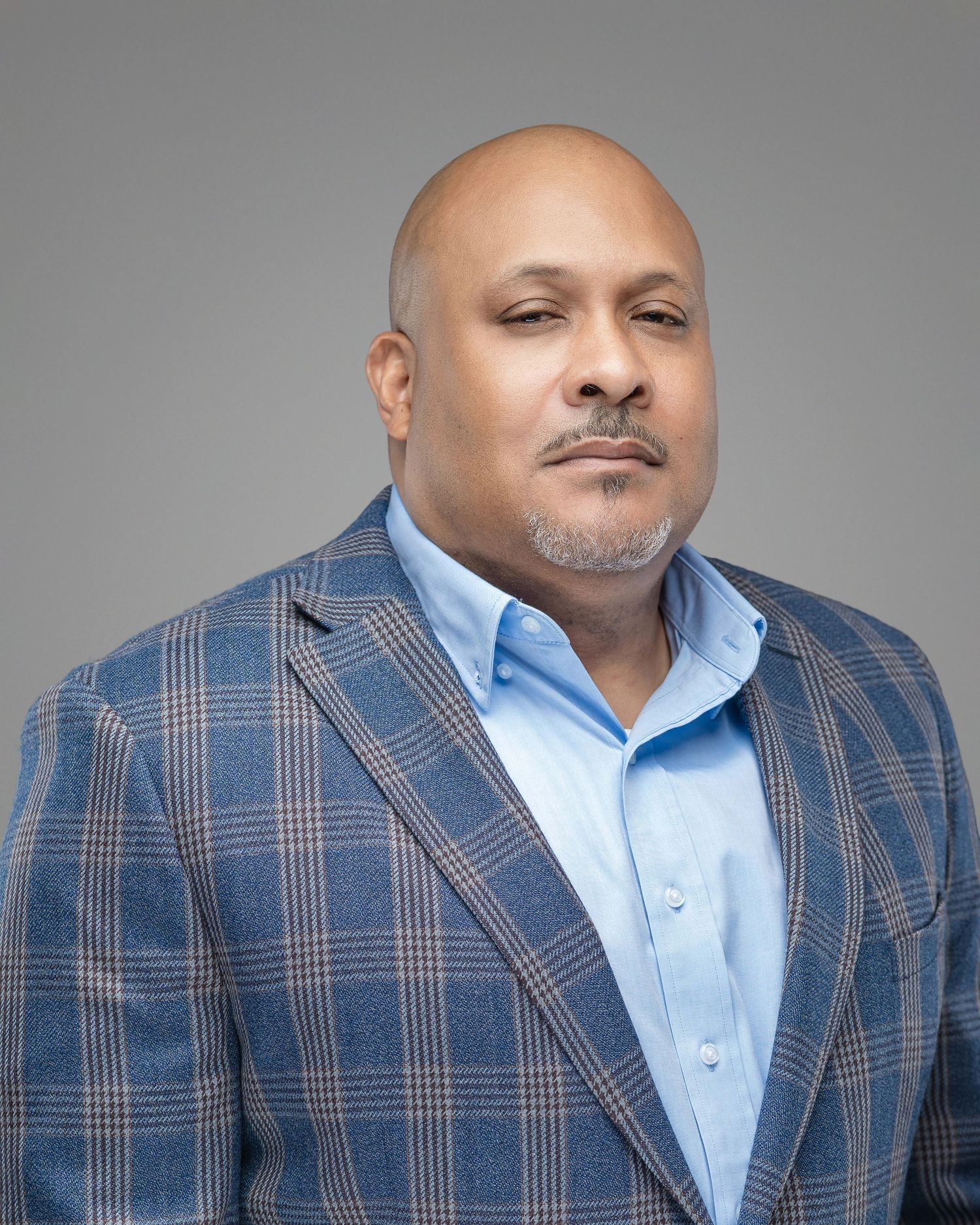 Professional headshot in blue plaid blazer and light blue shirt against gray background.