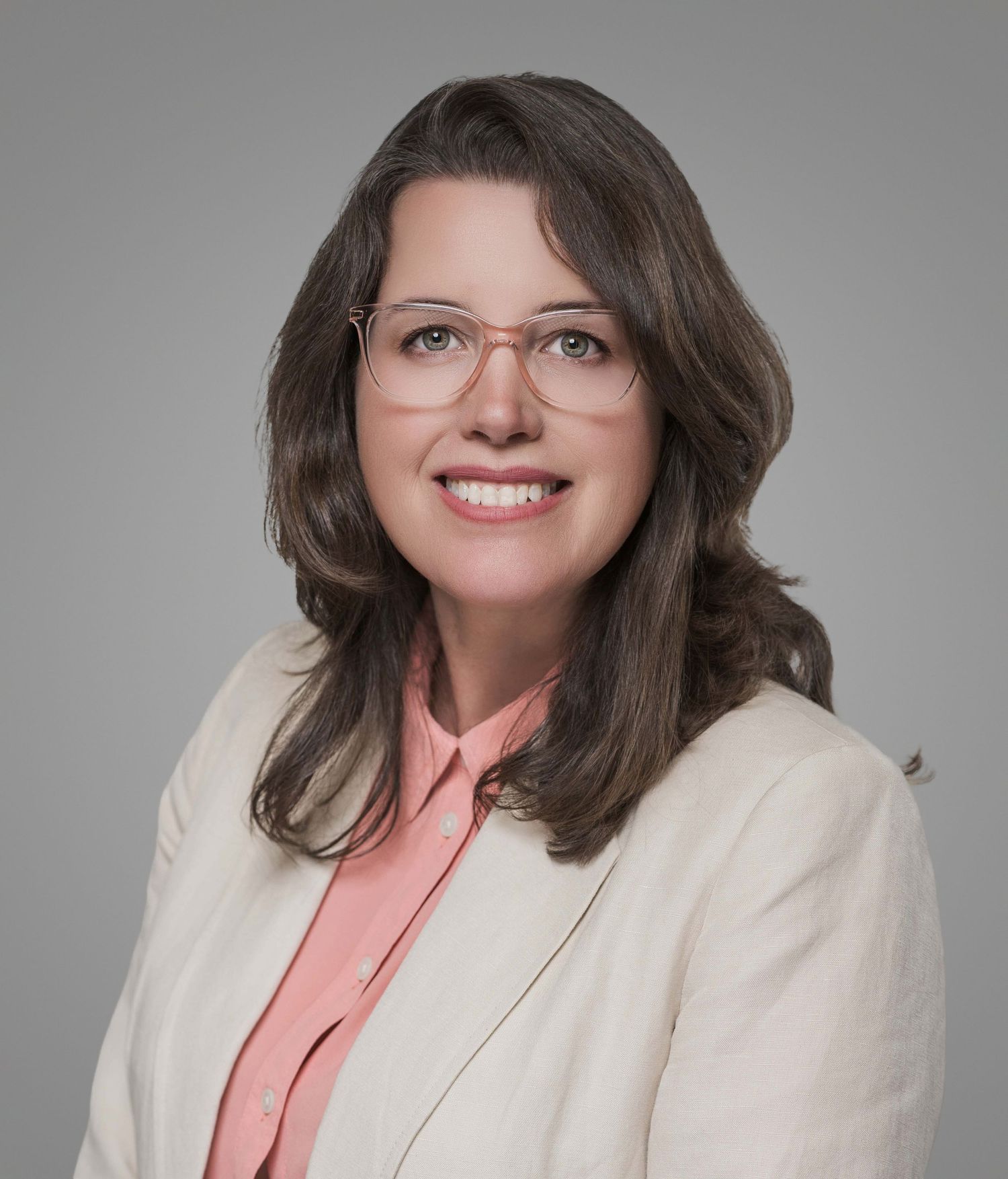 Professional headshot of a businesswoman in cream blazer and coral blouse wearing glasses.