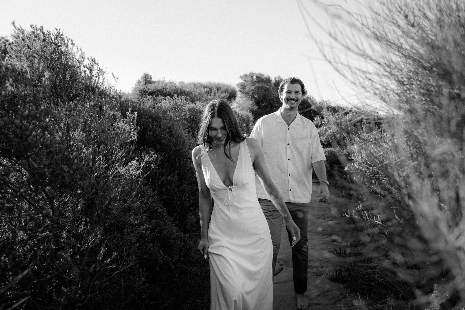 A couple in white clothing walking together on a winding path through natural foliage captured in black and white.
