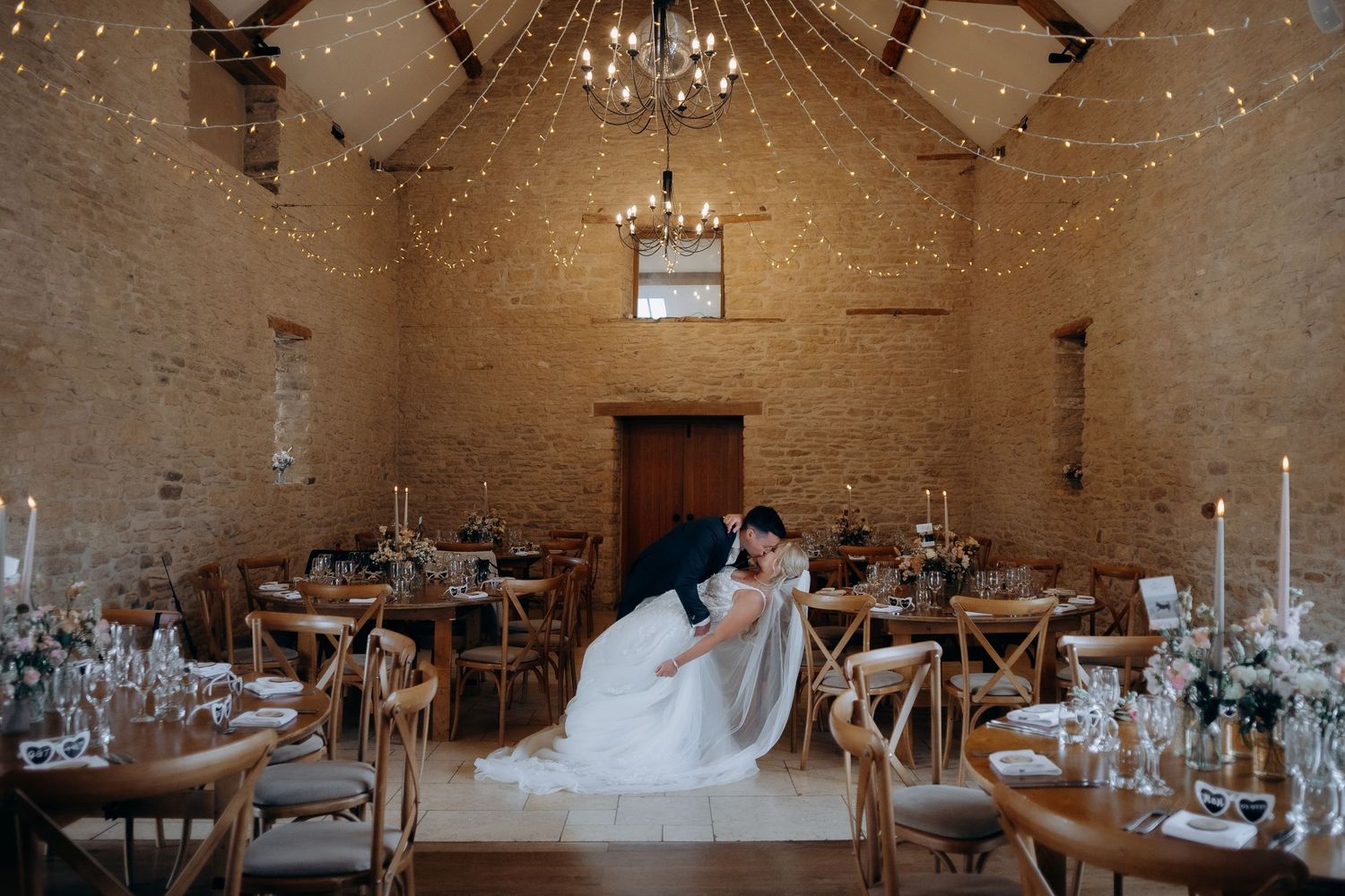 Inside a rustic stone barn venue with wooden tables and chairs set for a wedding reception with elegant candle centerpieces.