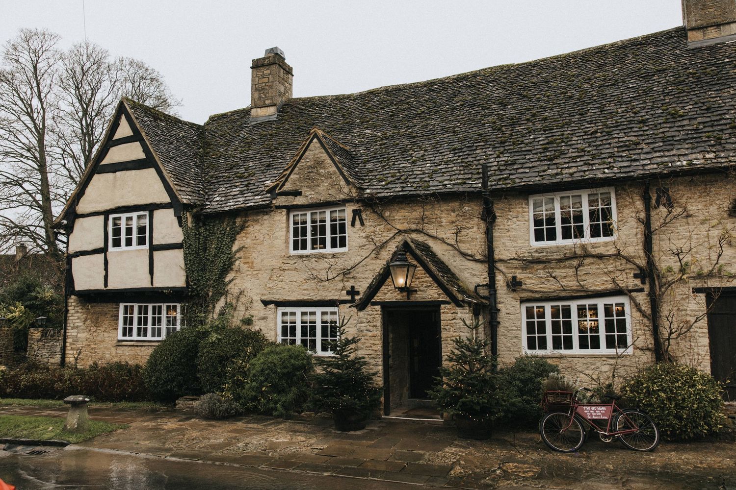 A charming Tudor-style cottage with stone walls and timber framing sits along a quiet village street with a bicycle parked outside.