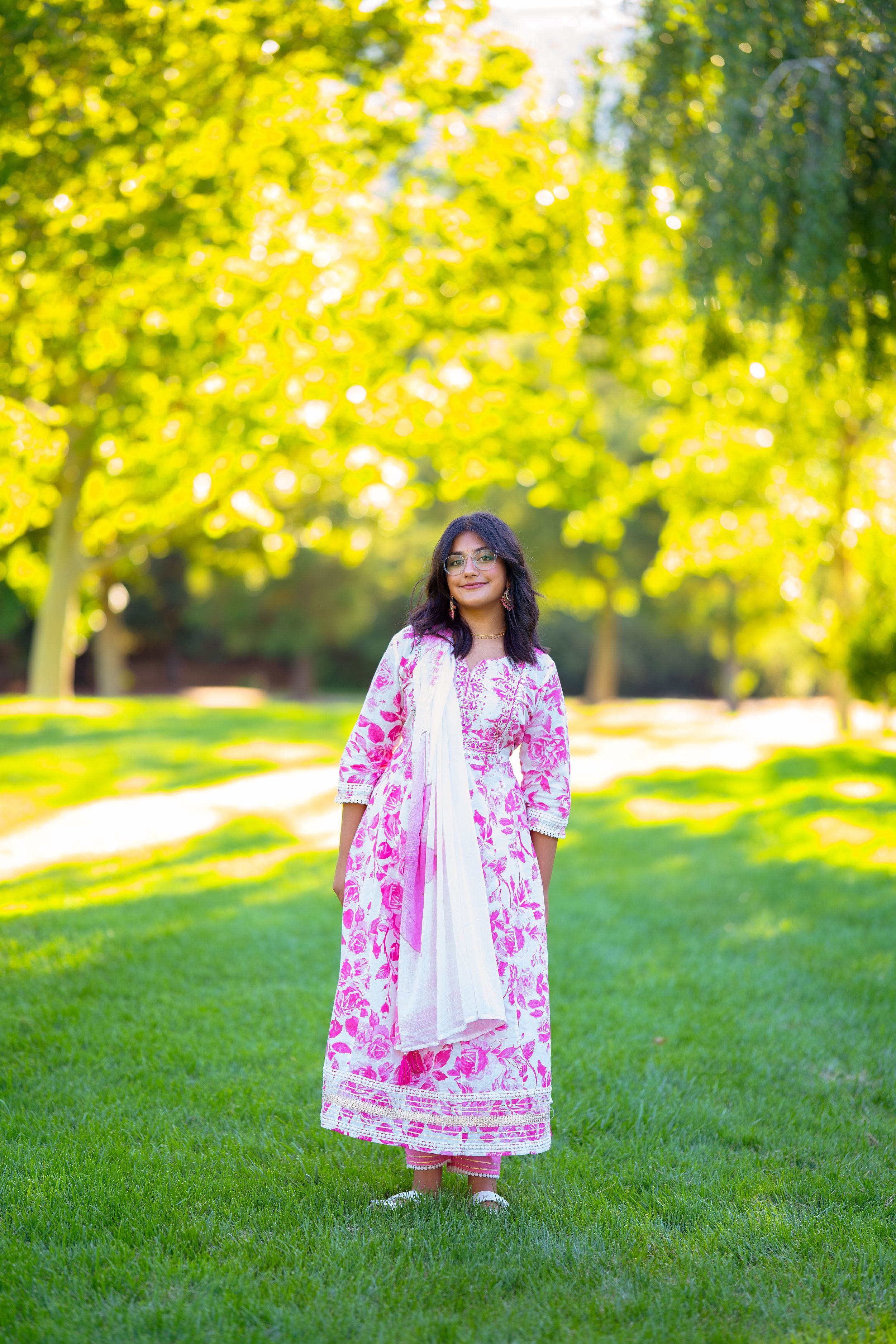 A person in a pink and white embroidered dress stands on green grass with sunlit trees in the background.