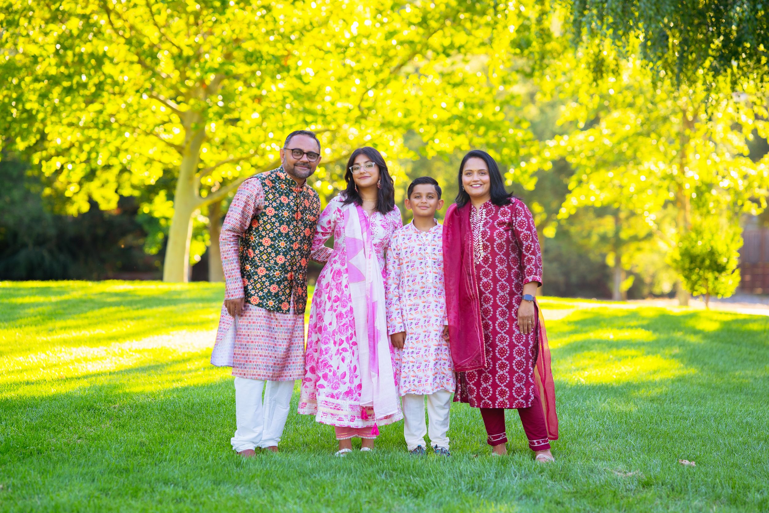 Colorfully dressed family in traditional South Asian attire poses together on bright green lawn with sunlit trees behind.