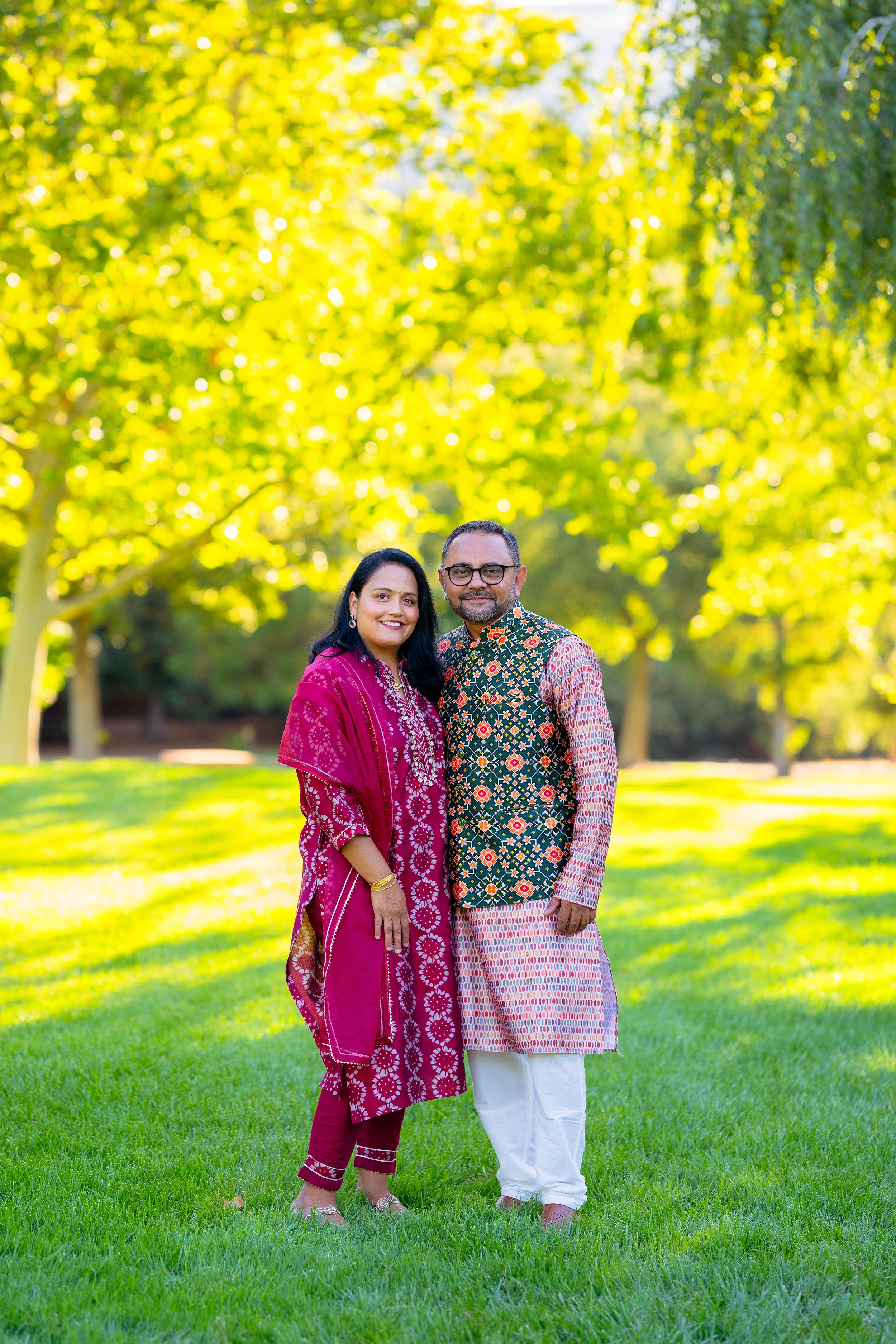 Two people wearing ornate traditional South Asian outfits pose together on grass with golden sunlit trees behind them.