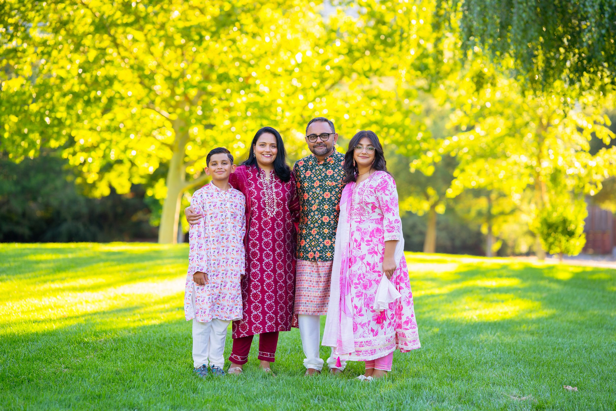 Group wearing coordinated pink, red and white holiday outfits stands on green lawn under golden fall foliage.