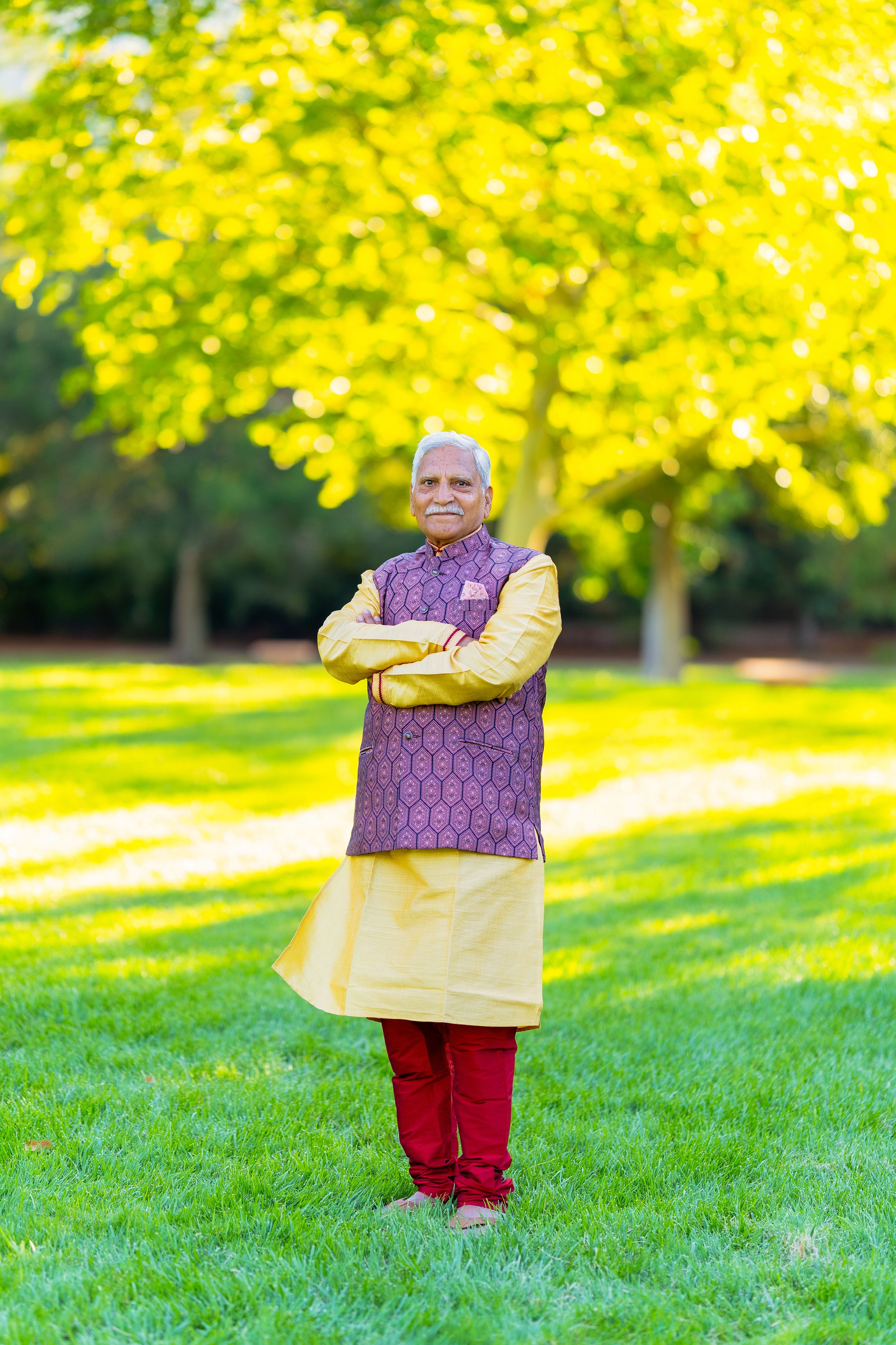 Someone in traditional Indian attire with a yellow kurta and purple vest stands in a park with vibrant autumn foliage.