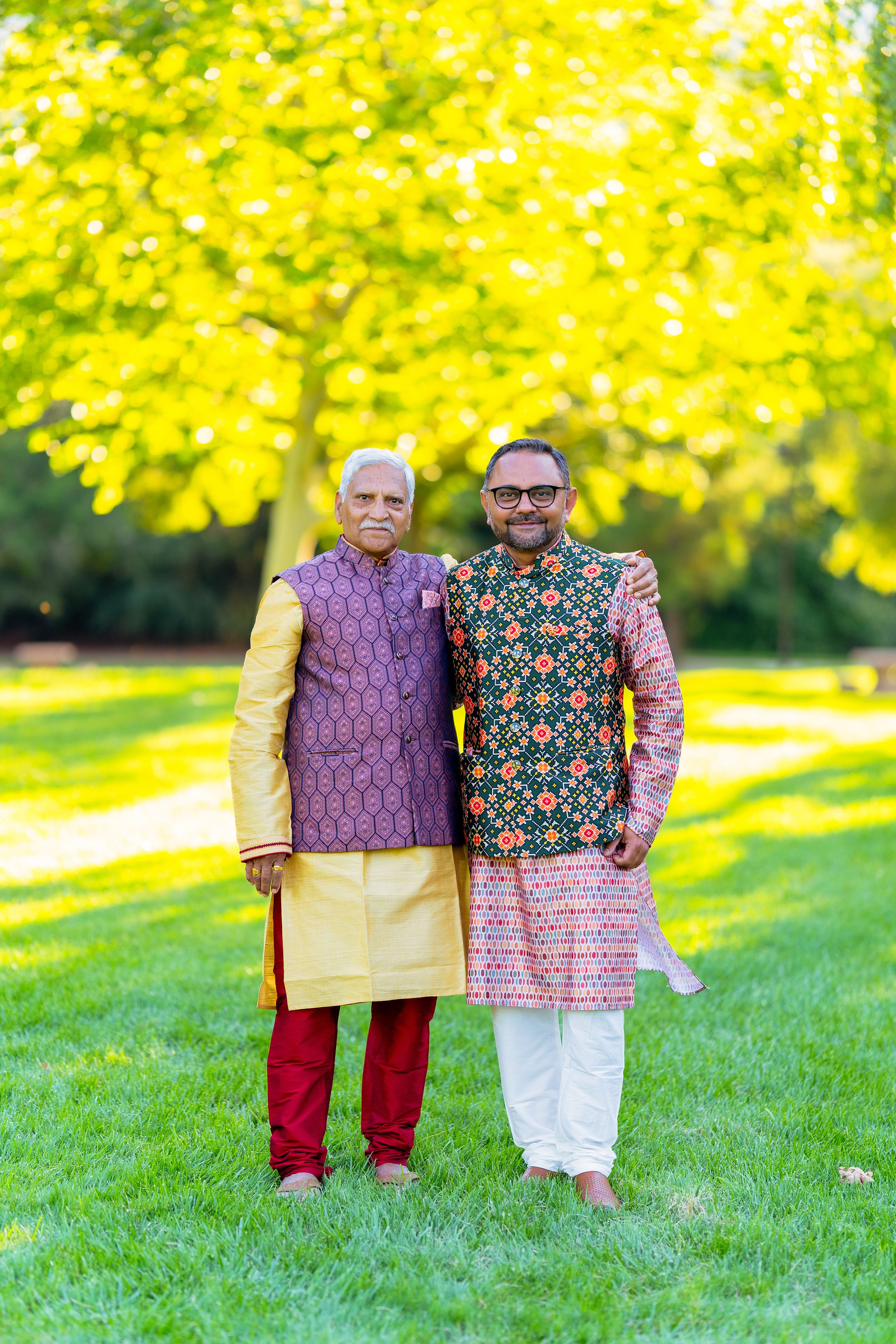 Two people in traditional Indian kurtas stand together on a lawn with vibrant yellow foliage in the background.