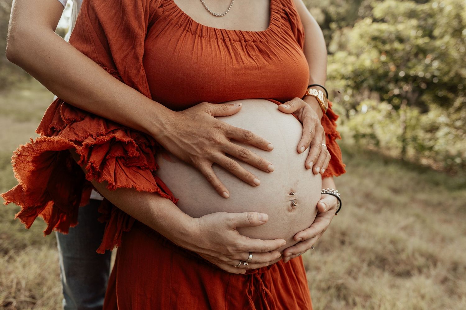A figure wearing a flowing red dress cradles their pregnant belly in a sun-dappled outdoor setting during sunset.