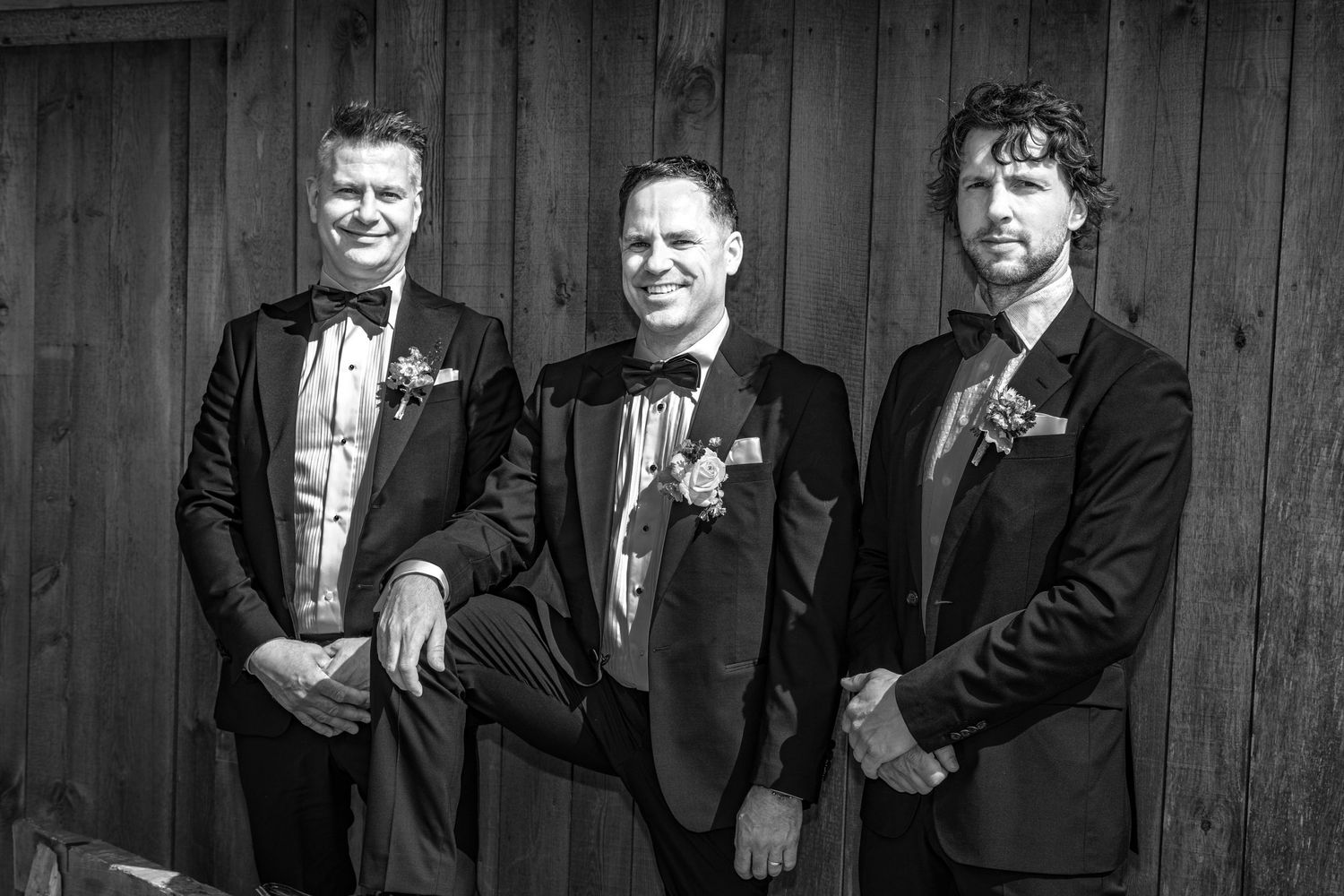 Three groomsmen in black tuxedos with boutonnieres pose together against rustic wooden barn wall.