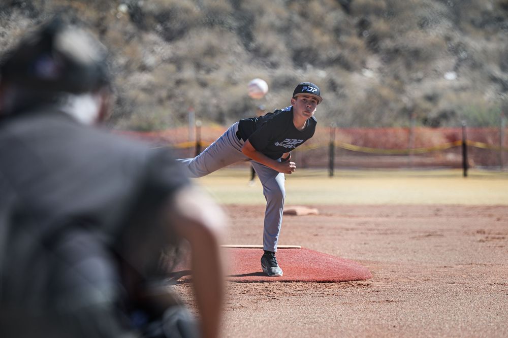 Youth baseball pitcher delivers a pitch toward home plate during a sunny outdoor game.