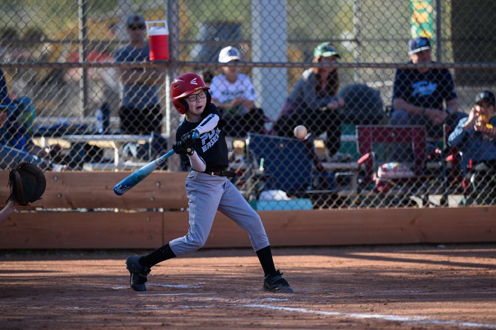 Young baseball player in red helmet swings at a pitch during a youth league game outdoors.