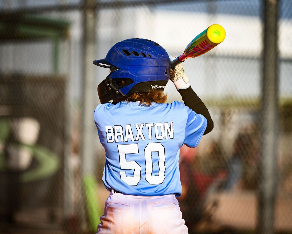 Young baseball player Braxton number 50 in light blue uniform stands ready to bat.