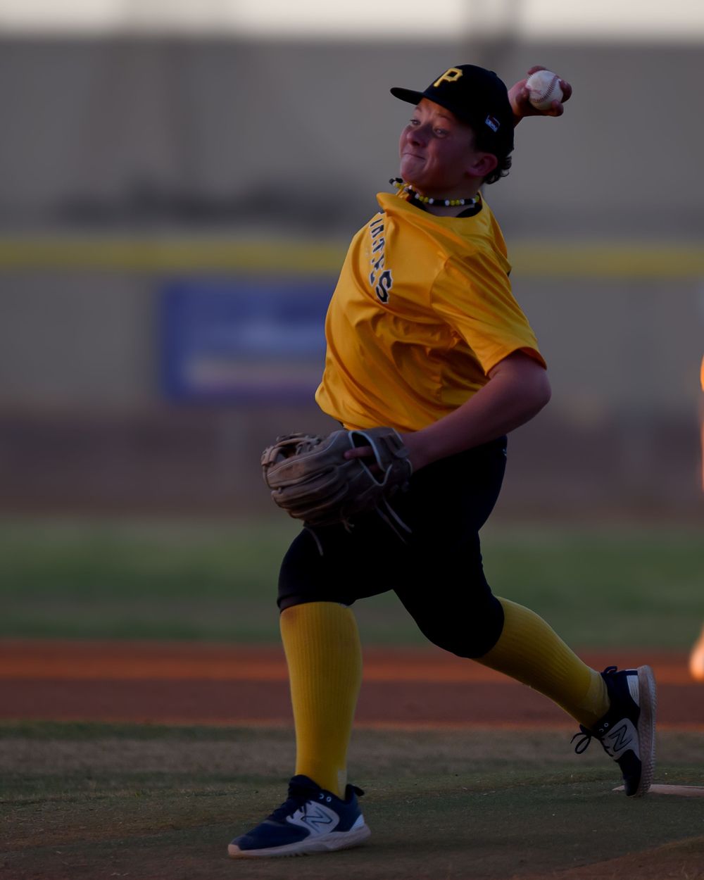 Youth Pirates pitcher winds up to throw in yellow uniform and black cap during an evening softball game.