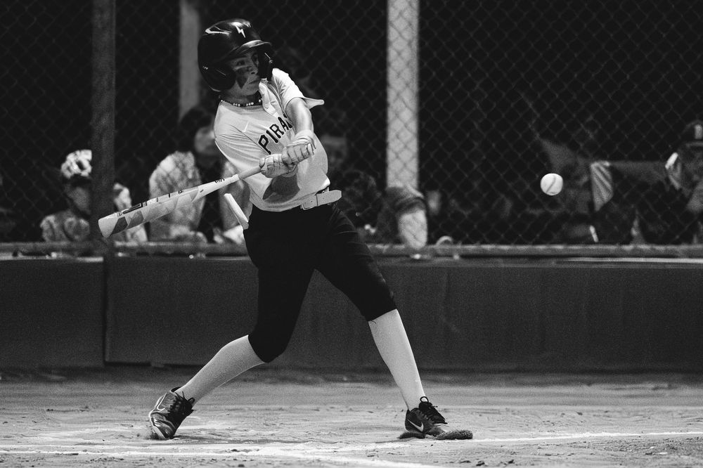 Black and white action shot of youth softball batter making contact with the ball at night.